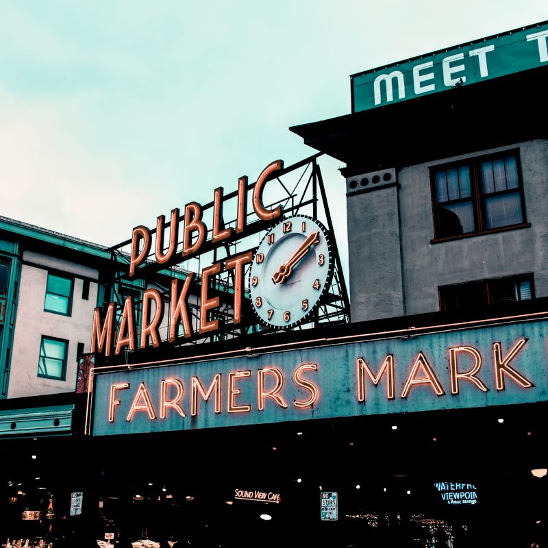 The image shows a historic neon sign for a public farmers market, set against an urban building backdrop under an overcast sky.