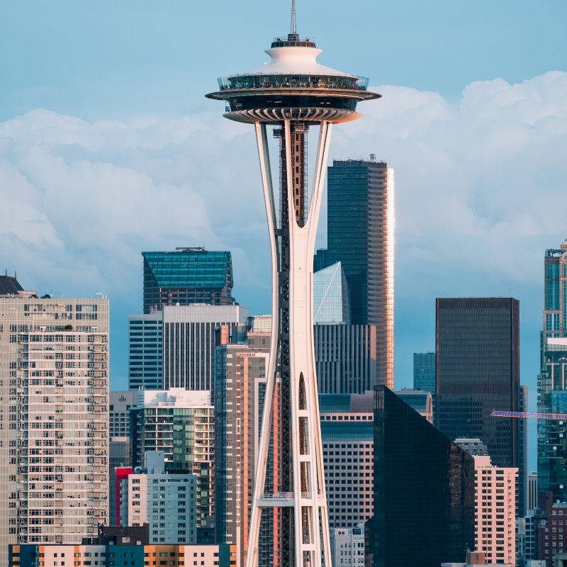 The image shows the Space Needle with Seattle's skyline in the background, featuring various tall buildings against a partly cloudy sky.