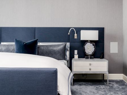 A modern bedroom with a blue and gray color scheme, featuring a bed, pillows, lamp, and a white nightstand with a decorative frame.
