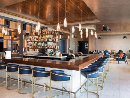 The image shows a sleek bar with blue stools, modern lighting, and well-stocked shelves, set in a spacious, contemporary room.