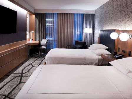 The image shows a hotel room with two beds, a wall-mounted TV, a desk, and large window with curtains, softly lit by lamps.