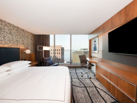 A modern hotel room with a bed, desk, chair, large TV, and city view window.