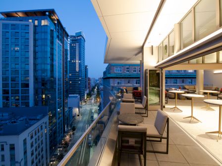 The image shows a modern rooftop terrace with seating, overlooking city buildings at dusk, featuring a blend of urban and architectural design.