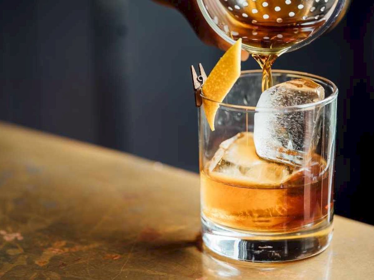 A cocktail being poured through a strainer into a glass with ice and a citrus twist garnish on the counter.