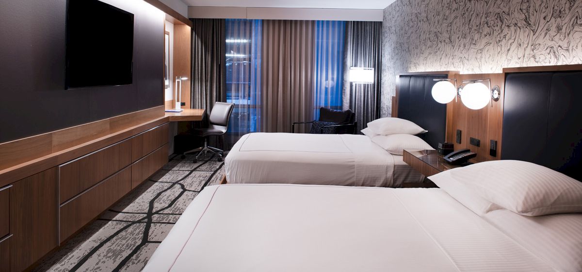 The image shows a hotel room with two beds, a TV on the wall, a desk, and lamps by the beds, in a modern, softly lit setting.