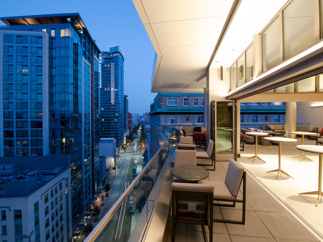 A modern rooftop terrace with seating overlooks a city's urban skyline at dusk, featuring tall buildings and a well-lit street below.