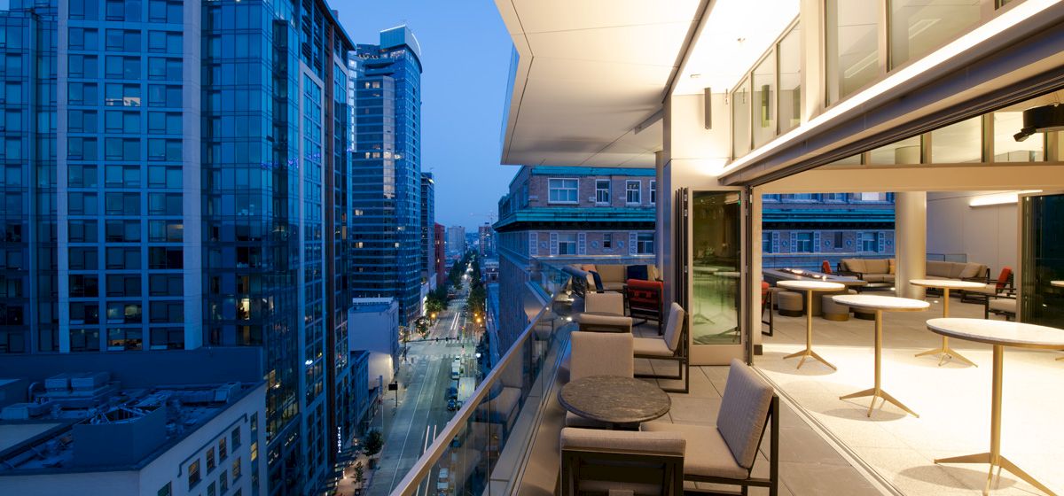 A modern rooftop terrace with seating overlooks a city's urban skyline at dusk, featuring tall buildings and a well-lit street below.