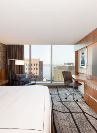 The image shows a modern hotel room with a large bed, a flat-screen TV, a desk, and a chair, featuring a view of the city through a window.