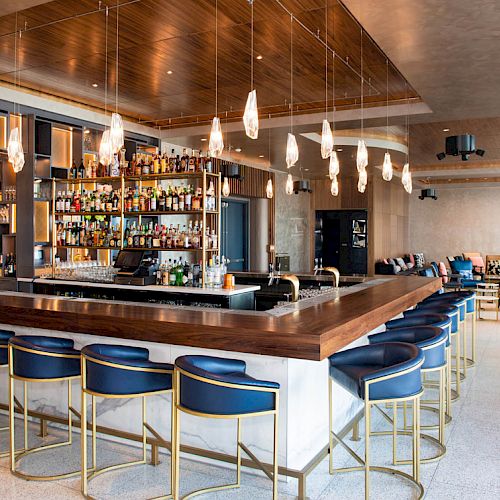 A modern bar with a central counter, surrounding blue stools, shelves of bottles, and a lounge area in the background.