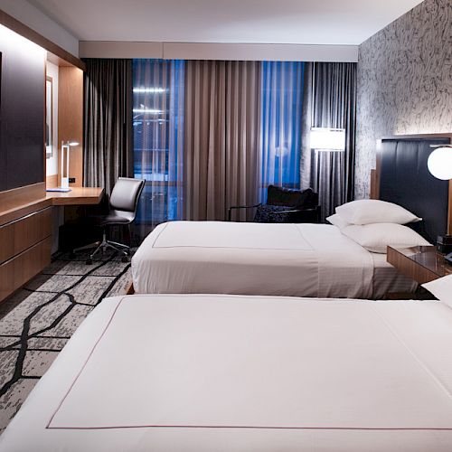 The image shows a hotel room with two beds, a desk, a TV, lamps, and curtains. The decor is modern and neutral in style.