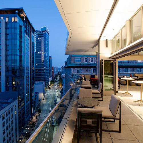 The image shows a modern rooftop terrace with seating, overlooking a cityscape of tall buildings at dusk.