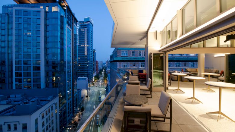 A modern cityscape at dusk with tall buildings and a lit rooftop patio featuring tables and chairs, overlooking a bustling street.
