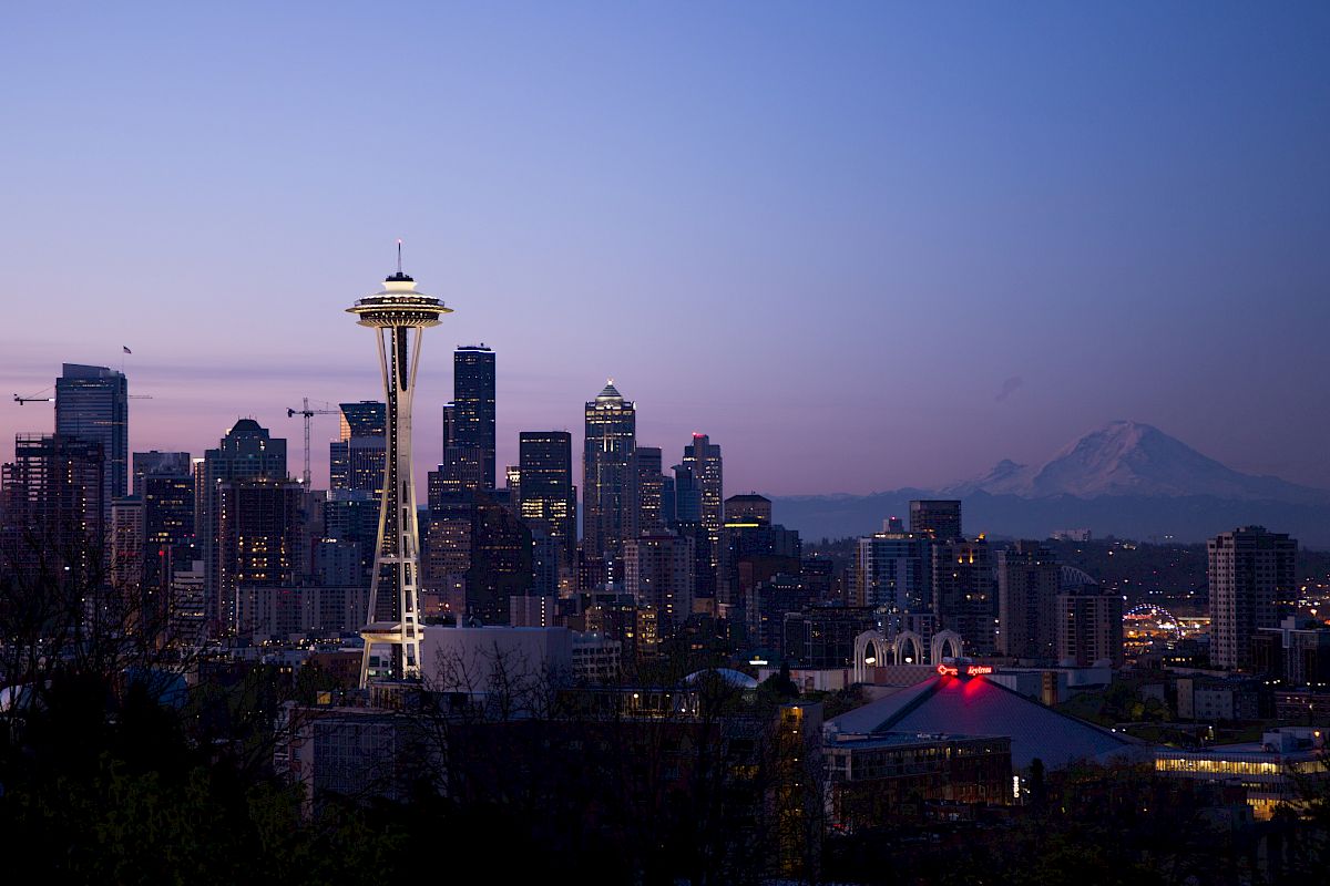 The image shows a skyline view of Seattle at dusk, featuring the Space Needle with distant mountains in the background.