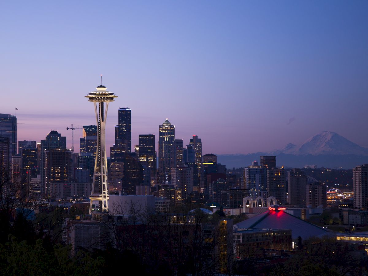 The image shows a skyline view of Seattle at dusk, featuring the Space Needle with distant mountains in the background.