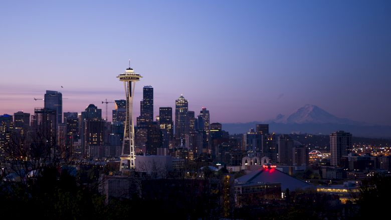 The image shows a skyline view of Seattle at dusk, featuring the Space Needle with distant mountains in the background.