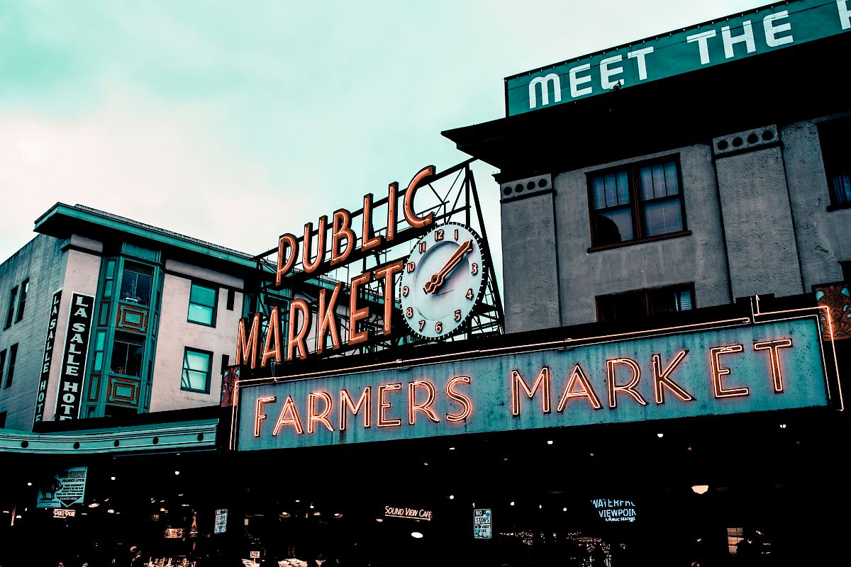 The image shows the iconic neon signage of a public farmers market with a large clock and surrounding buildings in the background.