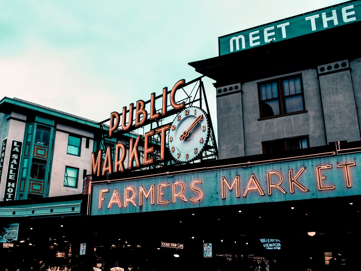 The image shows the iconic neon signage of a public farmers market with a large clock and surrounding buildings in the background.