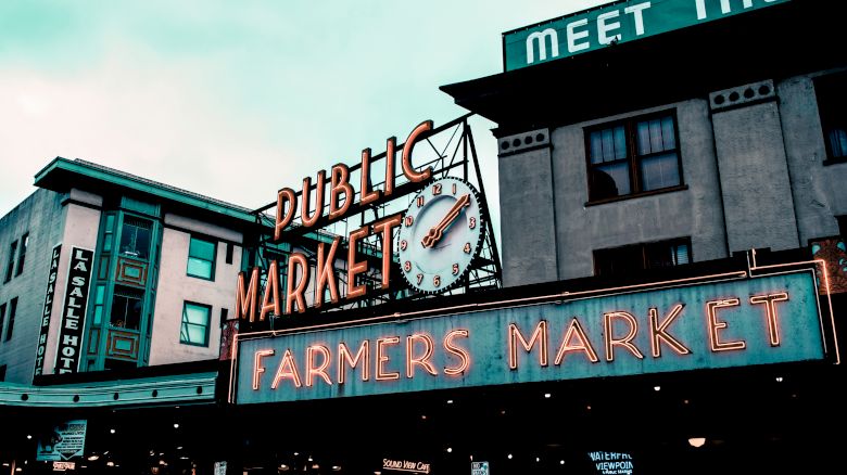 The image shows the iconic neon signage of a public farmers market with a large clock and surrounding buildings in the background.