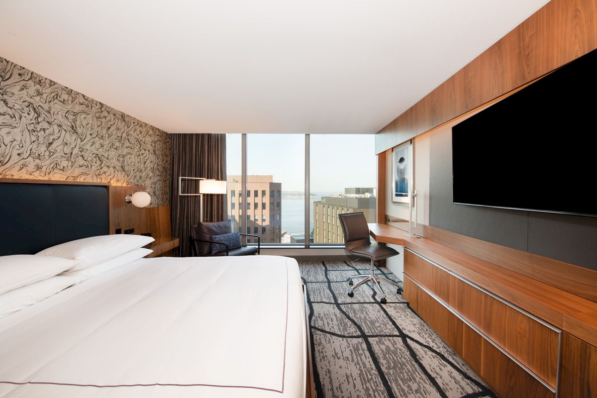 The image shows a modern hotel room with a large bed, wall-mounted TV, desk, and window with a city view.