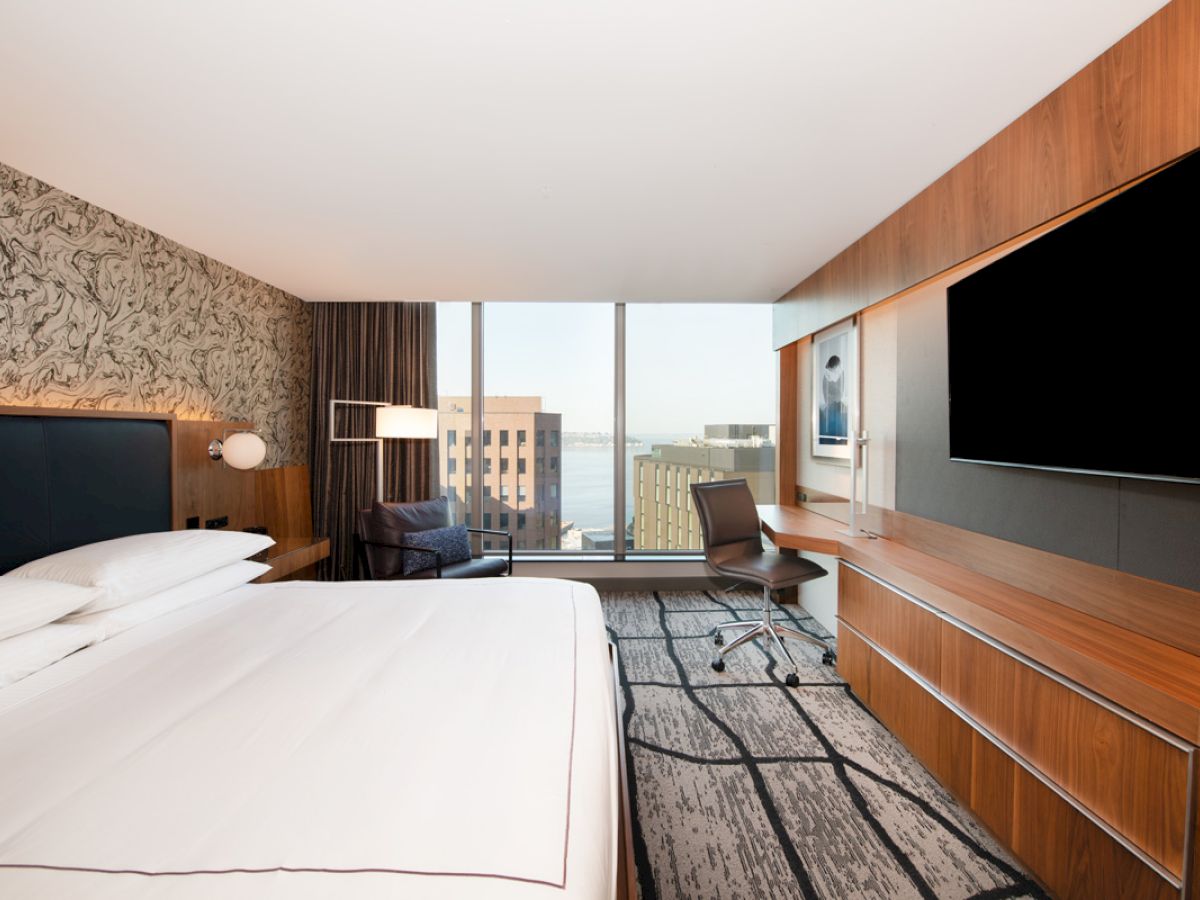The image shows a modern hotel room with a large bed, wall-mounted TV, desk, and window with a city view.