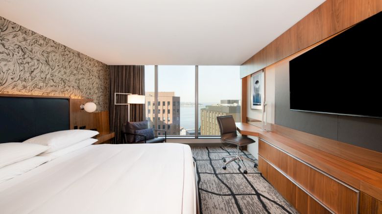 The image shows a modern hotel room with a large bed, wall-mounted TV, desk, and window with a city view.