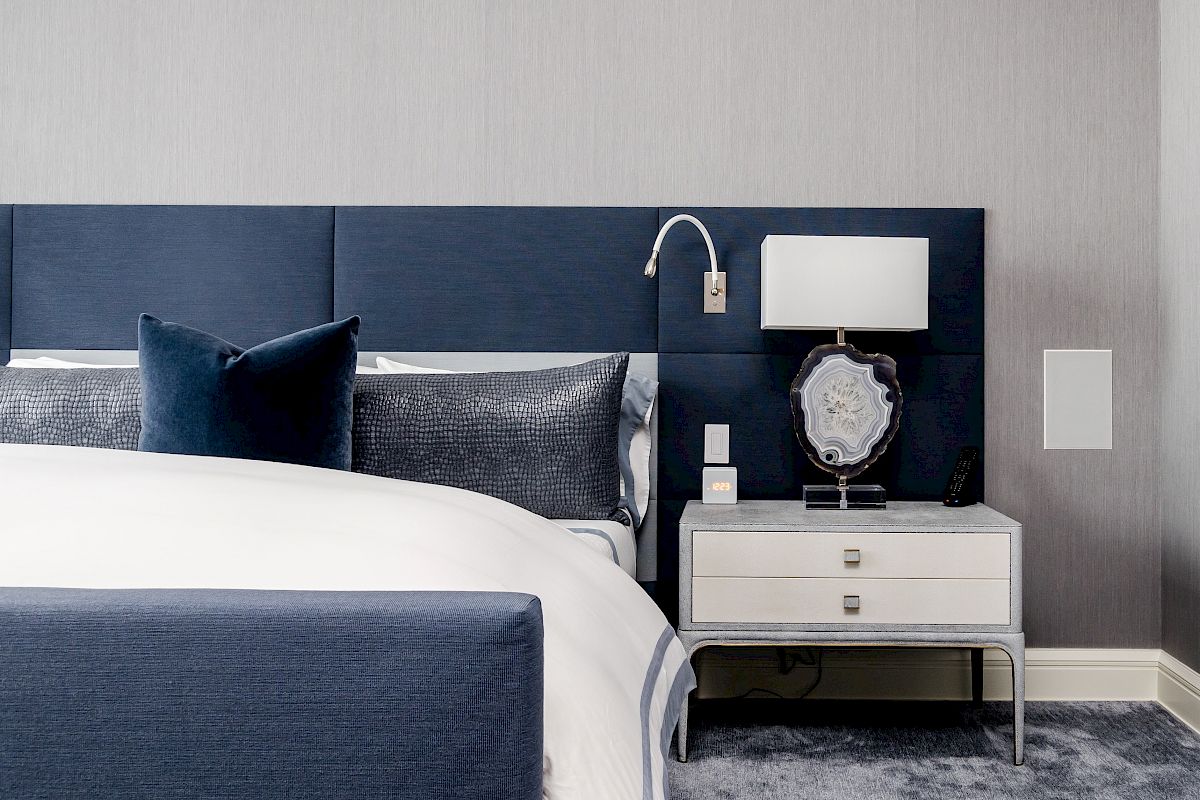 A modern bedroom with a navy bed, gray bedding, a white nightstand, a lamp, and wall decor. The design is sleek and contemporary.