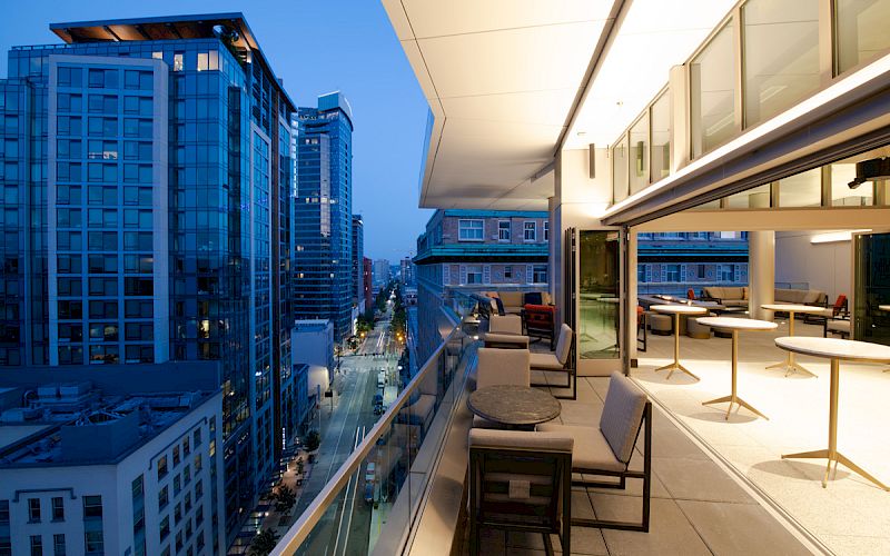 A modern rooftop terrace with seating overlooks a vibrant cityscape at twilight, featuring lit buildings and a clear sky.