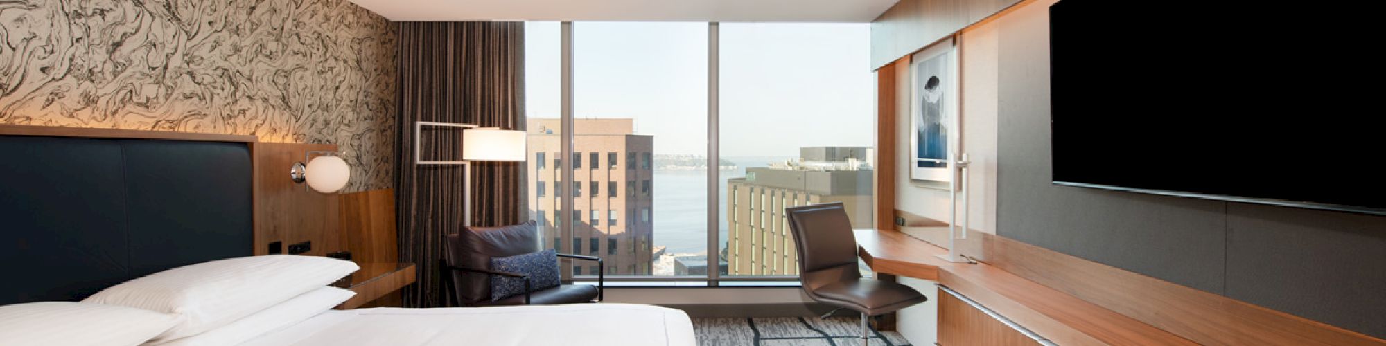 A modern hotel room with a large bed, TV, desk, and city view through a window.